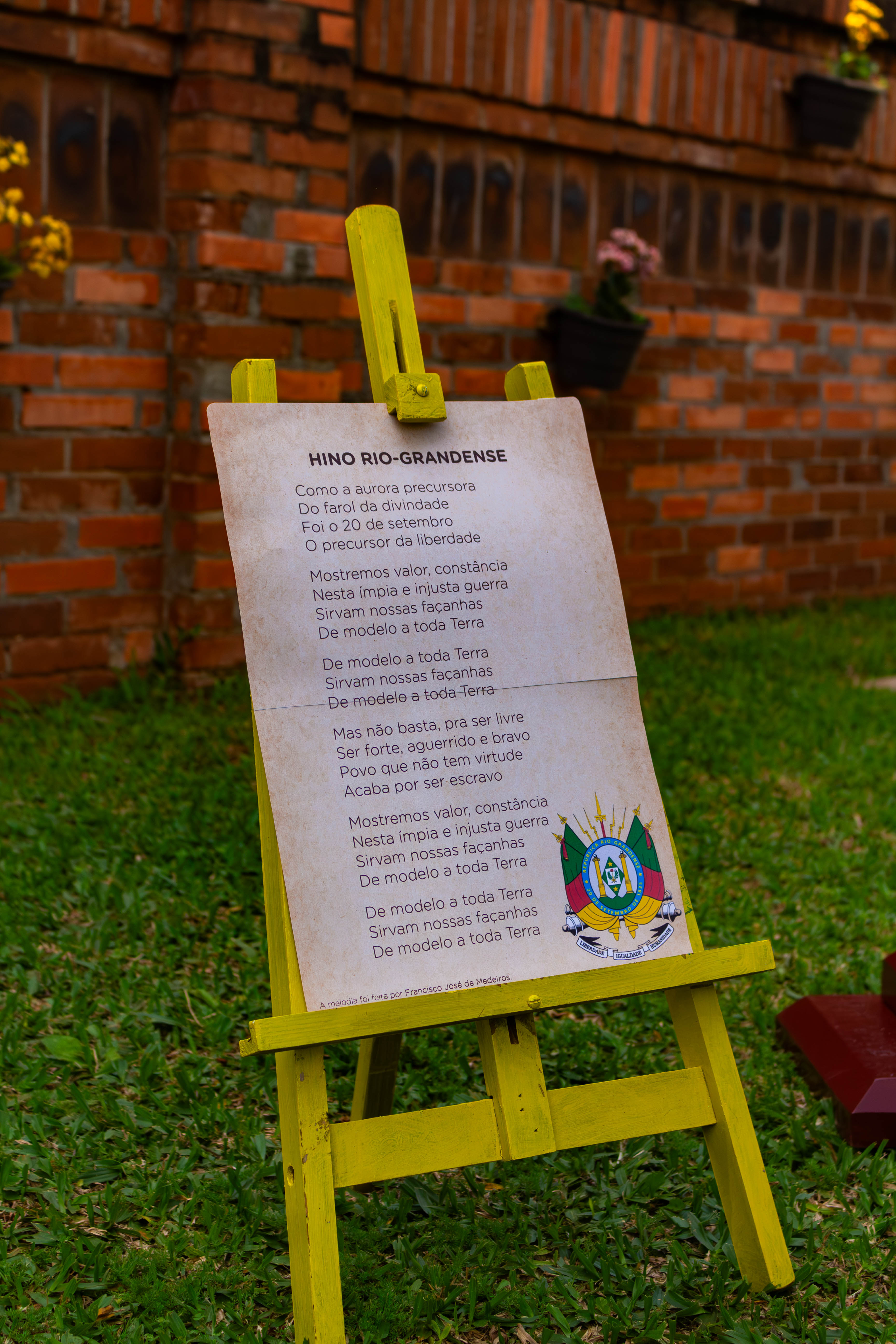 Letra do Hino Rio-Grandense impressa e apoiada em um cavalete de madeira amarelo ao ar livre